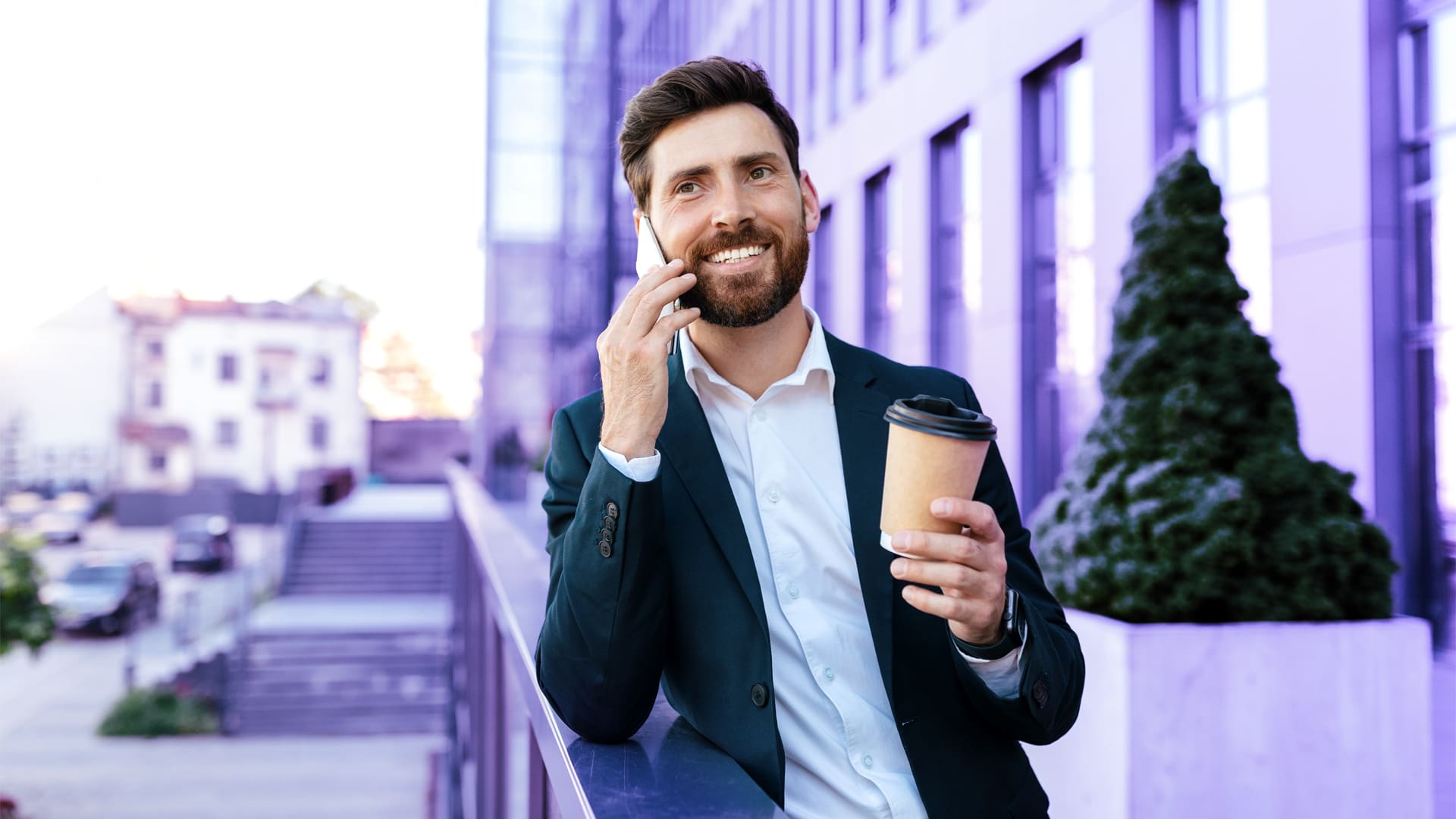 Hombre empresario sonriendo mientras va por la calle hablando por teléfono móvil con un café en la mano