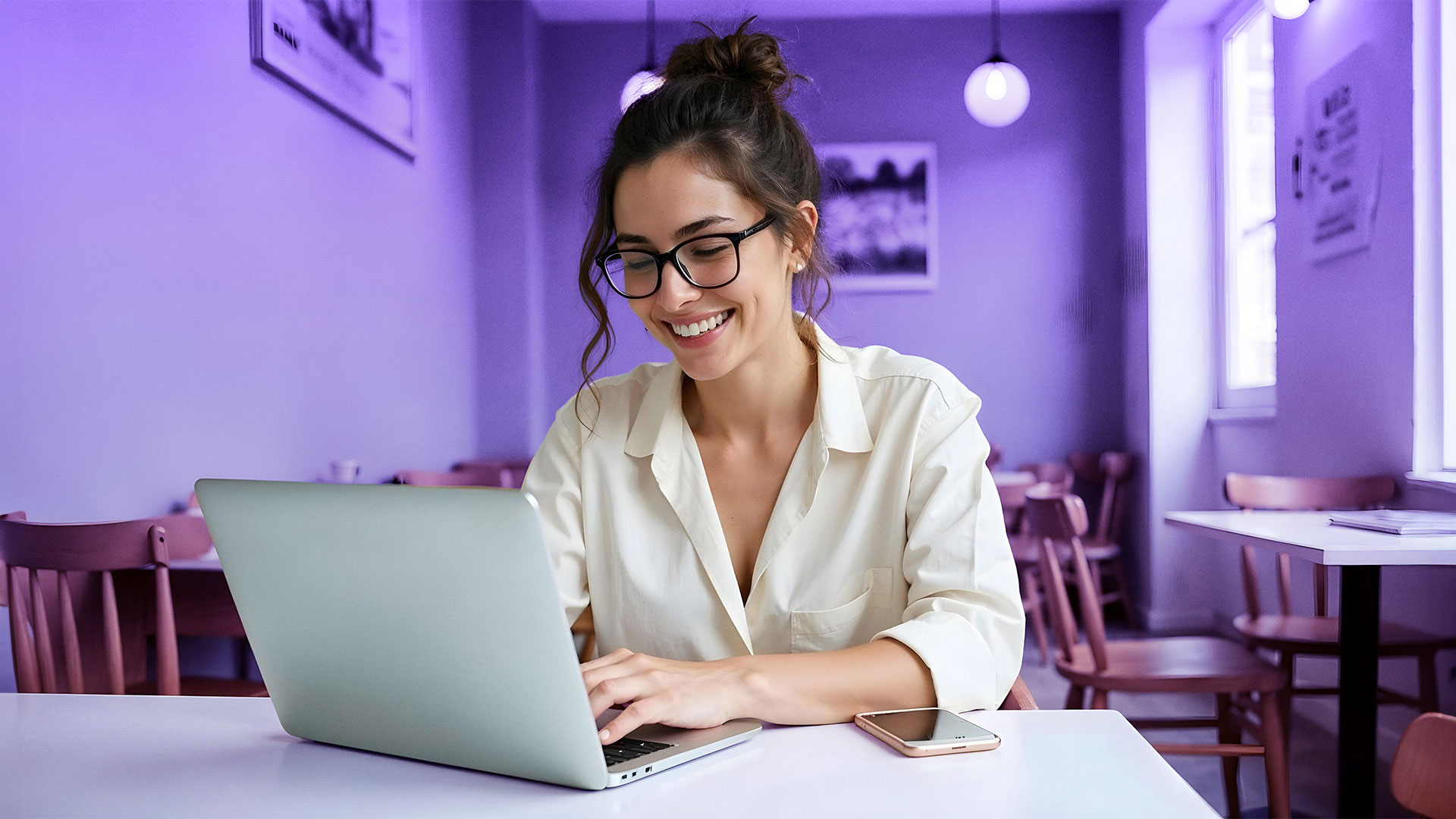 Mujer joven escribiendo por Microsoft Teams a su equipo desde el portátil de su empresa en un bar con el teléfono móvil al lado